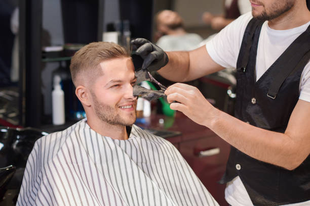 Smiling man sitting on chair in barber shop while barber in protective gloves removing black mask from face of client. Handsome man doing cleaning procedure against dots. Concept of care.