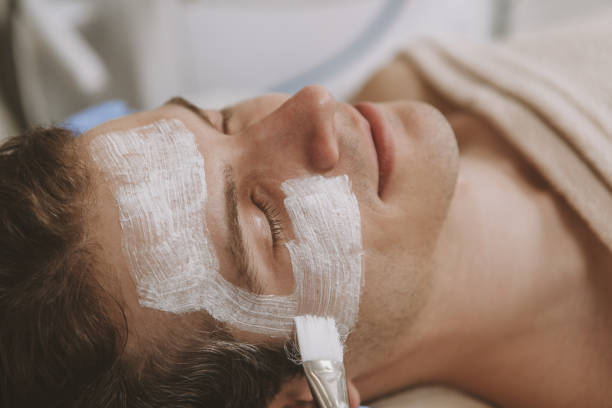 Cropped close up of a handsome relaxed man getting face mask by professional cosmetologist at beauty salon. Attractive male client resting at spa center, therapist applying face mask